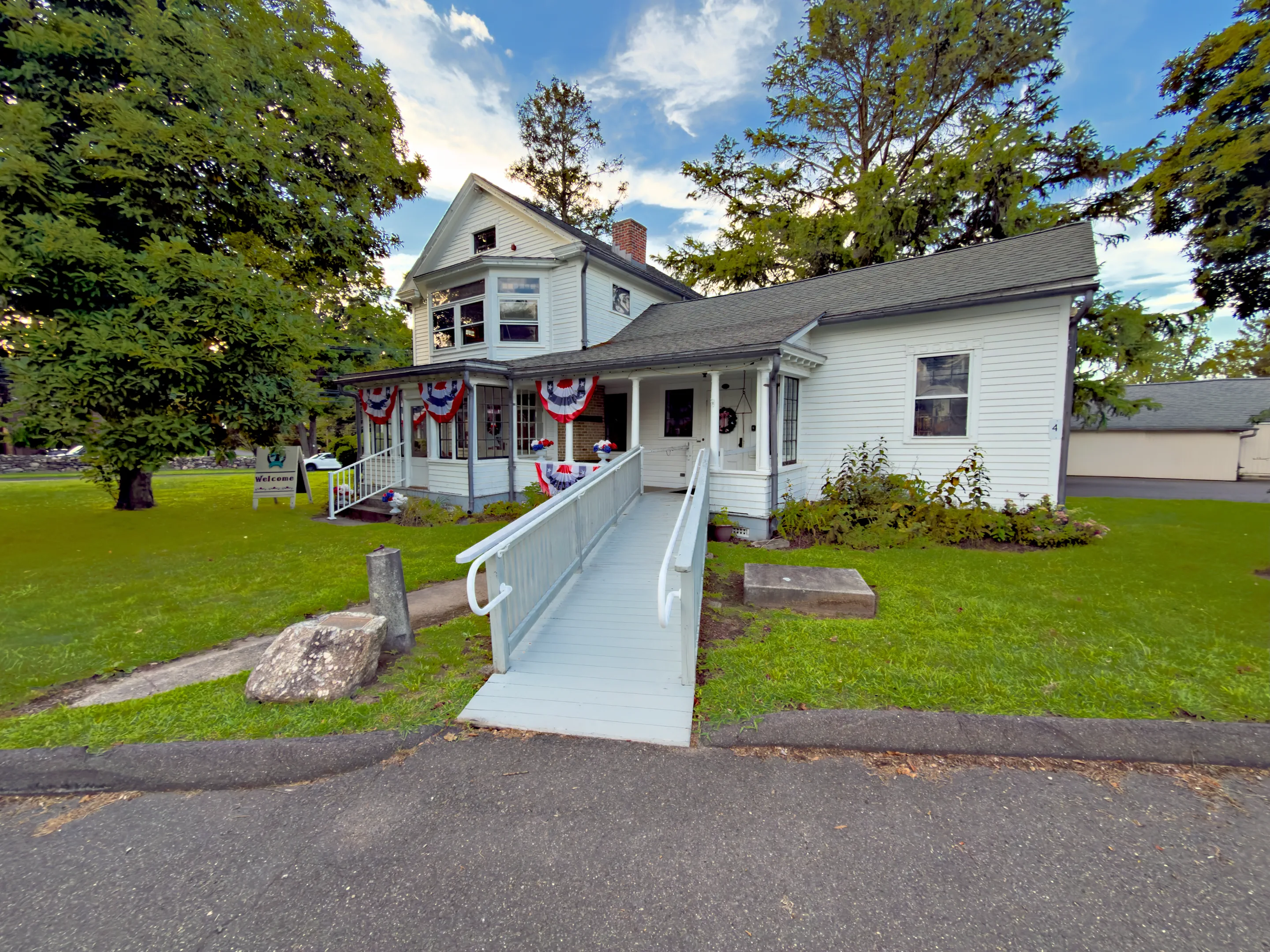 The white clapboard Boothe Homestead with flag bunting on its porch, an accessible ramp, and tall summer trees