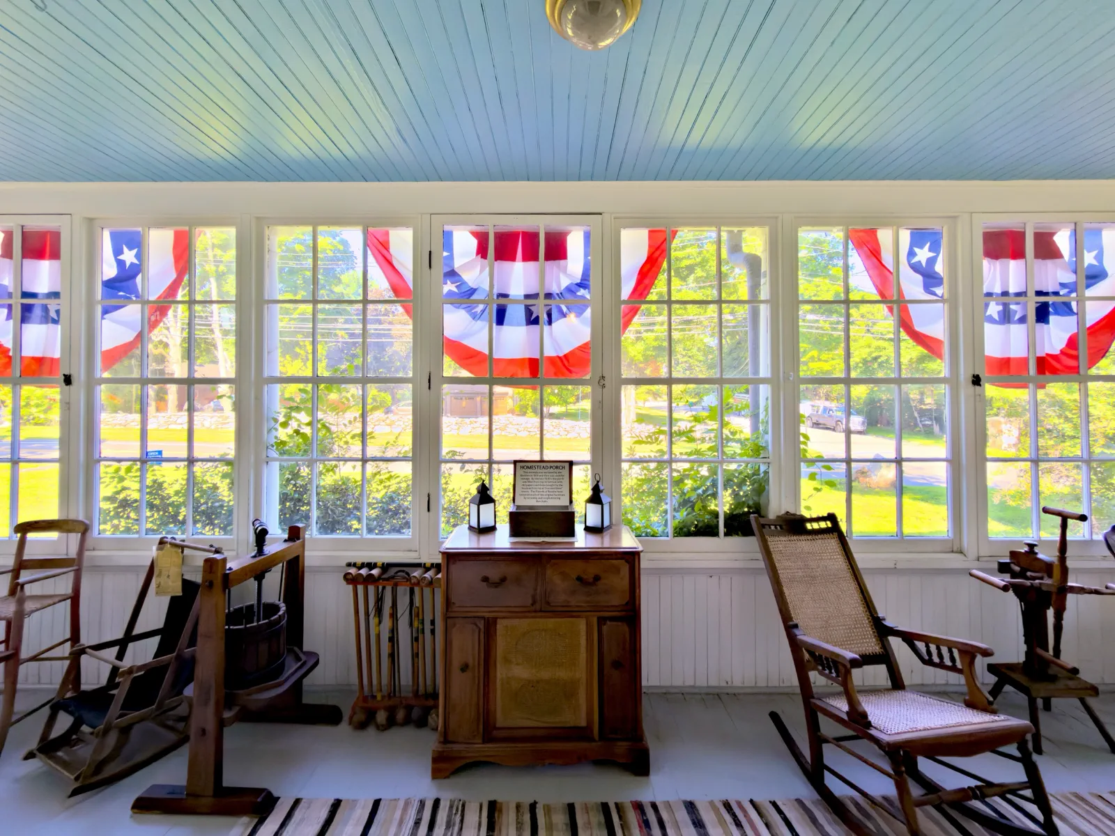 The enclosed Homestead porch with flag bunting in its windows, a cane rocking chair, a croquet set, and antique wooden furnishings
