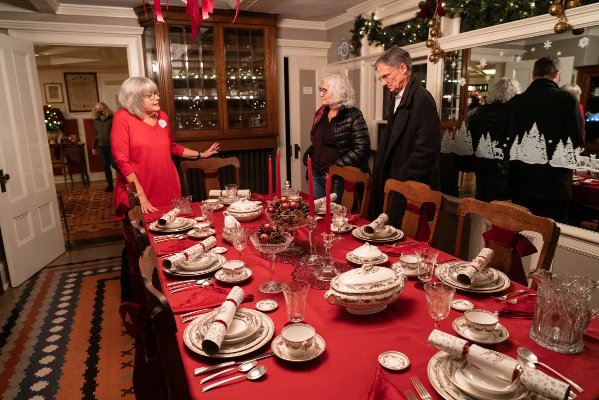 A Boothe Park volunteer speaking with visitors beside a holiday table setting in the Homestead