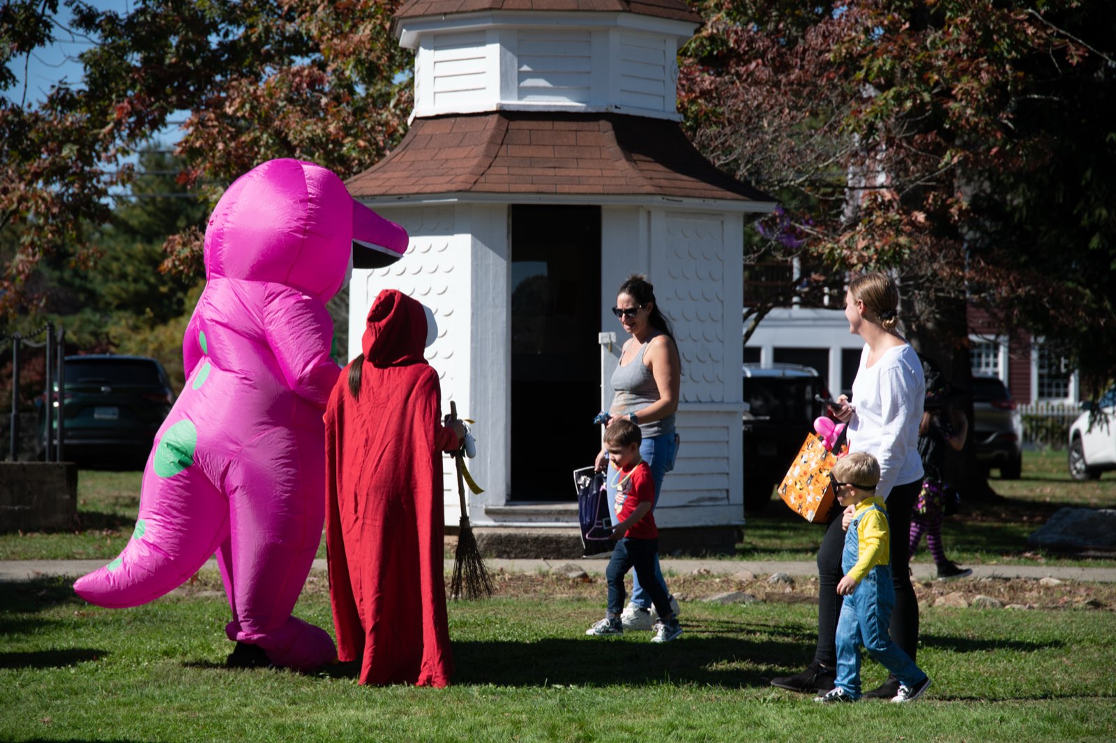 Visitors in costume, including an inflatable pink dinosaur, cross the lawn at Boothe Park